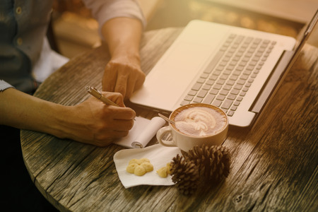 A man writes a pen in a notebook, to work in the office, laptop on  table, a white mug of coffee.の写真素材