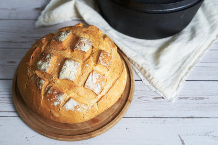 Round sourdough loaf showing its characteristic cuts on a wooden board, next to a cast iron pot and a tea towel, on a white wooden tableの写真素材