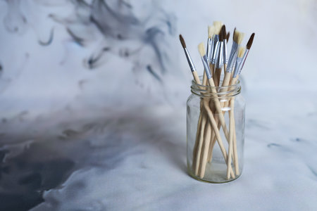 A collection of various paintbrushes with wooden handles displayed in a clear glass jar. The soft, abstract background creates a creative, artistic atmosphere.の写真素材