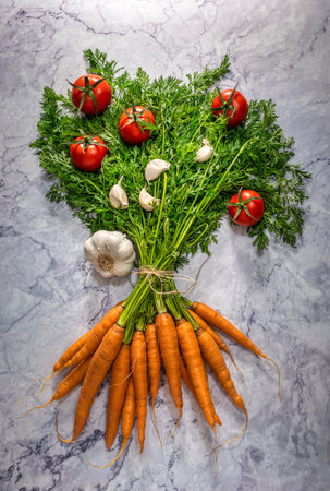 Raw carrots with green tops garnished with cherry tomatoes and garlic segments White marble...