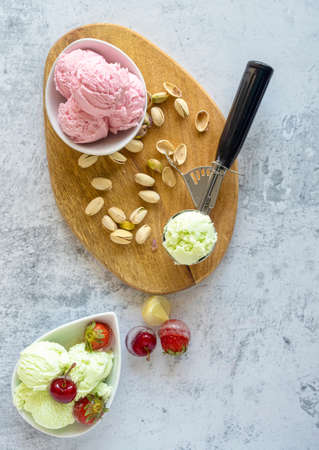 Bowl and scoops with delicious raspberry, cherry and pistachio ice cream.Wooden plate with delicious ice cream and different ingredients on the table decorated with branches and flowers.White stone background.の写真素材