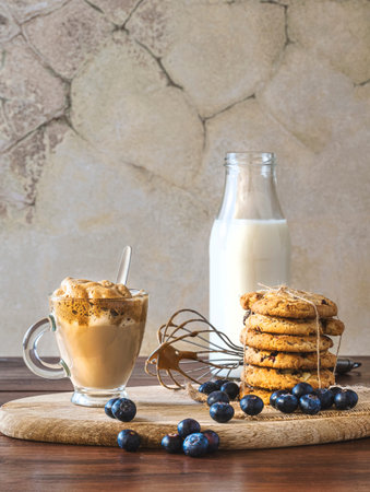 Dalgona coffee in a clear glass with blueberry cookies and blueberries on a wooden board. Stainless...