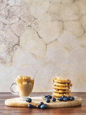 Dalgona coffee in a clear glass with blueberry cookies and blueberries on a wooden board. Rustic...