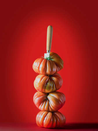 Close-up of ripe tomatoes placed one on top of the other with a knife creating a perfectly balanced tower. Red background. Beefsteak tomatoes.の写真素材