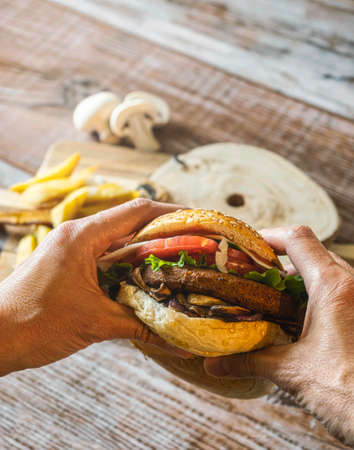 Hamburger with chickpea patty, tomato, mushrooms and vegetables on rye bread. Two hands hold the burger. On the bottom fries and mushrooms. Copy space.の写真素材
