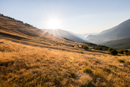 Sunrise on top of the mountain with a view of the nearby mountain range and a lake in the background. Yellow grass in summer and green mountains in front, seen from the Marsicano mountain in Abruzzoの写真素材