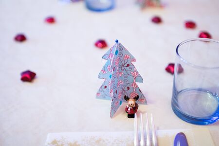 Christmas decorations with paper Christmas trees and a Reindeer mascot dressed as Santa Claus under a handmade paper tree. Decorations for a white table of Christmas dinner and lunch with red heartsの写真素材
