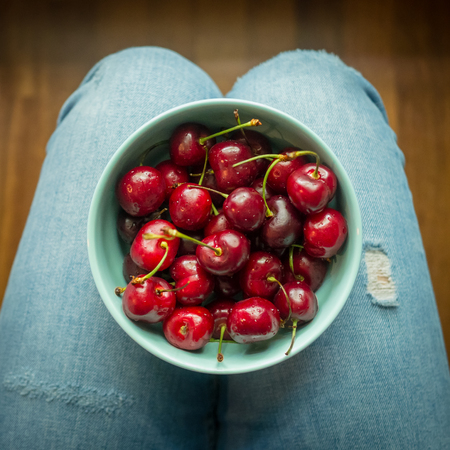 Vintage look image of a light blue bowl full of red cherries on woman's legs wearing jeans. Squared format. Top view.の写真素材