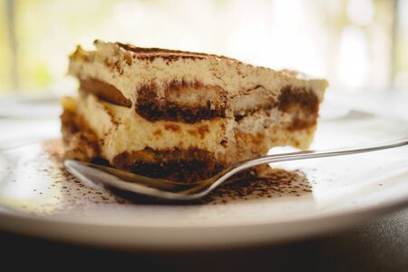 Close up of a slice of homemade tiramisu (traditional Italian dessert) on a white plate with a spoon on a marble board. Landscape format.の写真素材
