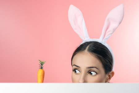 Beautiful young woman with pink bunny ears and toy carrot on pink background. Minimal Easter concept.の写真素材