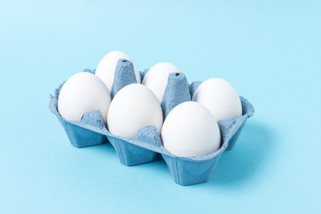 White eggs in carton tray on blue background. Minimal concept.