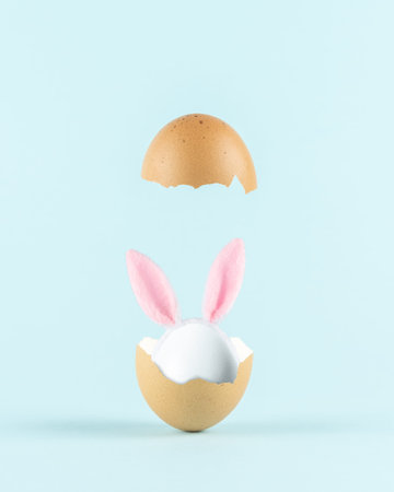 White egg with rabbit ears in cracked eggshell against pastel blue background. Easter minimal...
