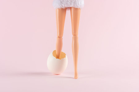 Girl doll leg in cracked Easter egg on pastel pink background. Easter minimal concept. Creative...