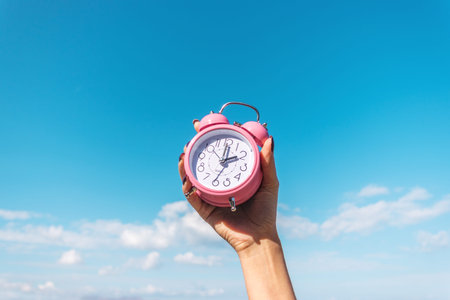 Woman's hand is holding pink alarm clock on the sky background. Summer vacation concept.