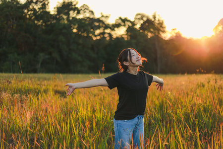 Portrait of a beautiful woman in black clothes rejoices in the green meadow Standing with arms wide spread Enjoy the beauty and tranquility of nature and the sunset light and bokeh.の写真素材