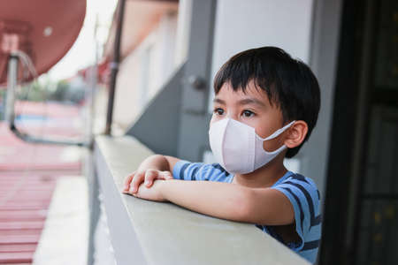 New normal The concept of staying at home Distance to stop the spread of covids 19 Portrait of a child wearing a white mask in a blue striped shirt looking out of the house to take care of health anの写真素材