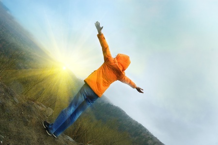 Young woman with backpack standing on cliff's edge and looking to a sky with raised handsの写真素材