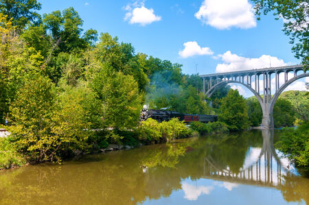 BRECKSVILLE, OH - SEPTEMBER 7, 2014: A steam locomotive on the Cuyahoga Valley Scenic Railroad pulls a passenger train under an arched bridge over the Cuyahoga River south of Cleveland as part of the annual Steam in the Valley event.のeditorial素材