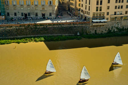 FLORENCE, ITALY - MAY 31, 2019: Three sailboats, seen from above, float past on the Arno River in the old section of Florence, with tourists and locals on the opposite bank.のeditorial素材