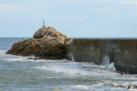 wave breaking on the wall of a harbor with a lighthouse at the end and a cloudy blue sky