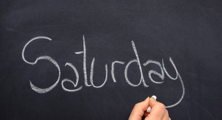 Woman's hand writing the day of the week on a blackboard with white chalk, Saturday