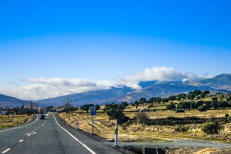 Road with road signs and snowy mountains with clouds around background with a nice blue skyの写真素材