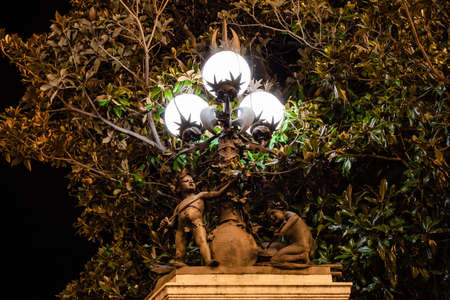 Madrid, Spain; 05/08/2020: Two bronze angels with a sword and playing the drum at the foot of a lamppost with three lights lit under the leaves of a tree next to the Buenavista Palace Gardensのeditorial素材