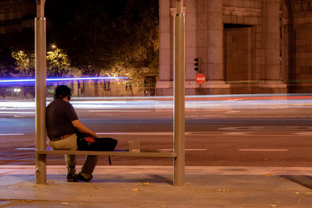 Worker waiting for the bus at a bus stop in Puerta de Alcala in Madrid while the vehicles draw colored lines in the air and on the asphaltの写真素材