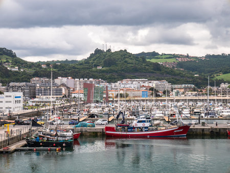 Laredo, Cantabria, Spain; 08-17-2021: Fishing boats docked in the fishing port of Laredo next to the building of the fishermen's brotherhood of San MartÃ­n with the town in the background a cloudy dayのeditorial素材