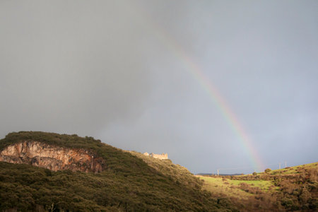 On a rocky mountain stands the castle illuminated by the only rays of the sun that break through after a storm and a large rainbow stands out in the skyの写真素材