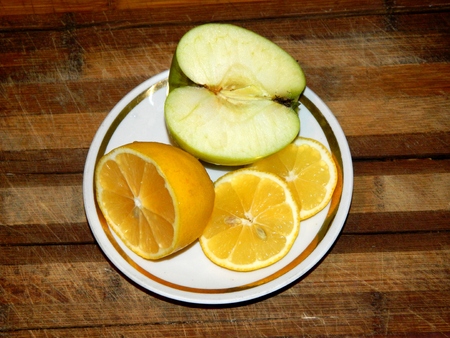 on a wooden chopping board in a white saucer with golden rim is half green apple and lemon and two slices of lemon cut on a horizontal surface in front viewの写真素材