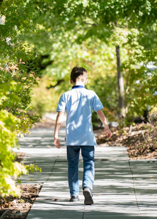 Blond teenage boy casual clothing walking relaxed along alley with trees and shadows. Bushes as blurred background.の写真素材