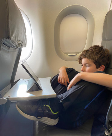 Adorable little boy in relaxed position traveling by plane. Child sitting by aircraft window and using a digital tabletの写真素材