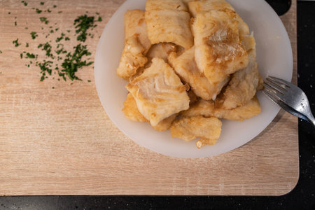 Pieces of fried hake loins over white plate and wooden cutting board. Preparing roman hake