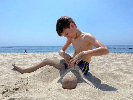 Child sitting on sandy beach pouring sand with his hands over his legsの写真素材