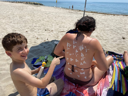 Smiling young boy applying sunscreen on his mother shoulder in the beach.