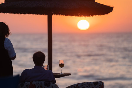 Rear view of a young romantic couple drinking cocktails and wine on a tropical beach at sunset on vacation or honeymoonの写真素材