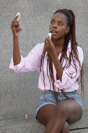 Portrait of young afro american beautiful girl putting on makeup sitting in urban streetの写真素材