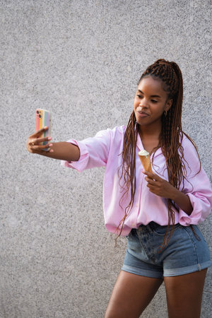Beautiful smiling black lady is enjoying an ice cream while taking a selfie isolated on grey...