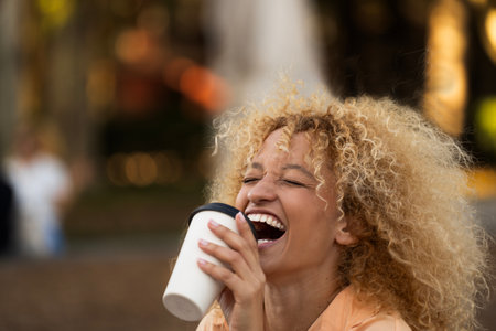 side view headshot of young latin woman with curly hair laughing while taking a coffeeの写真素材
