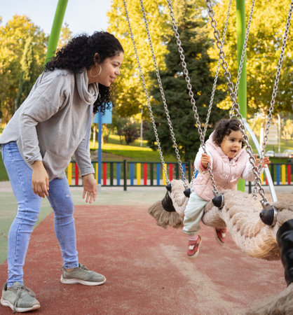 Smiling dominican mother and little kid girl having fun on a swing in a playground.の写真素材