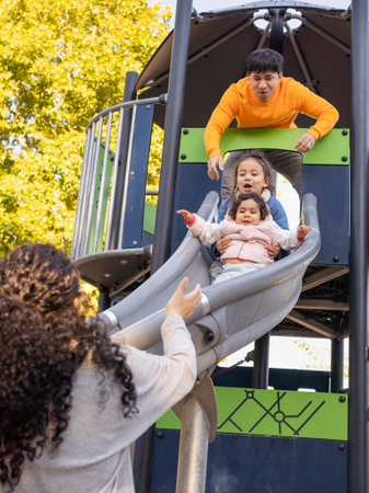 Hispanic father throwing their excited kids on a slide while mother waiting for them outdoors in a playground.の写真素材