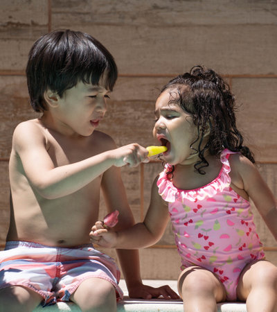 Two children sharing popsicles by the pool during a hot summer dayの写真素材