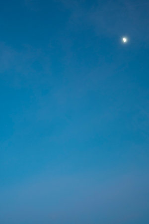 Clear blue sky with a visible crescent moon, creating a serene and unique daytime sceneの写真素材