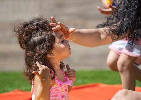 Mother carefully applies sunscreen to her young daughter's forehead, protecting her delicate skin...