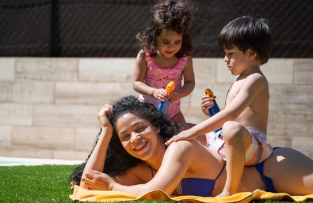 Latin mother relaxing and smiling by the pool, lying on a towel on grass, while her children are...