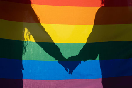Silhouettes of two people holding hands are projected on a rainbow flag, symbolizing lgbtq+ love, rights, and communityの写真素材