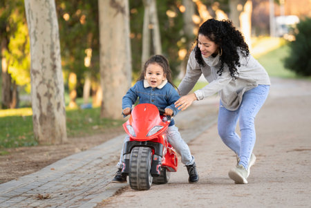 Mom helping her daughter riding a toy motorcycle in a parkの写真素材