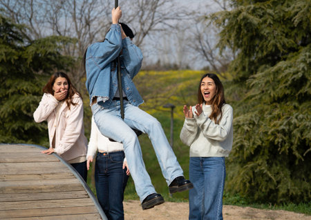Three young women enjoy a sunny day at the park, with one swinging on a zip line while her friends watch and cheer her onの写真素材