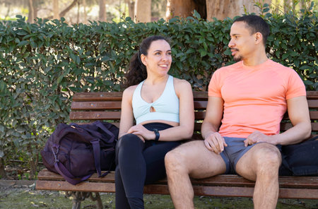 Smiling fit multiethnic couple resting on bench in park after training, talking and enjoying break with copy space.の写真素材